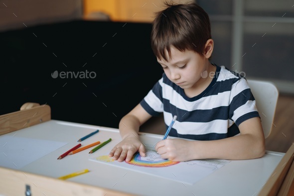 caucasian boy drawing a rainbow with pencils sitting at the desk in his room at home. - Stock Photo - Images