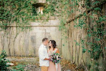 Groom hugs bride near the stone wall overgrown with green ivy