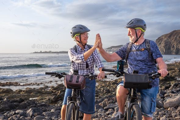 Happy senior caucasian couple on the pebble beach with electric bicycles at sunset give five smiling