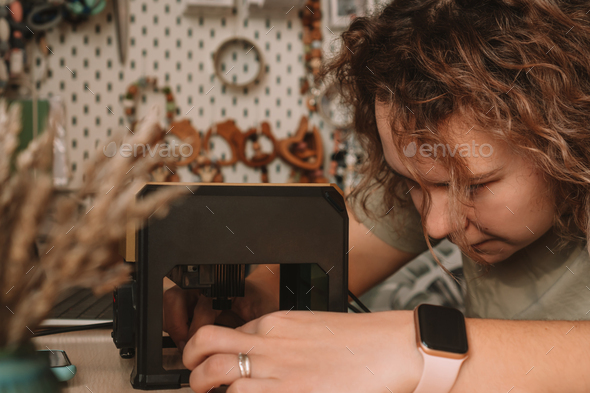Curly haired woman holds item in desktop laser wood burning machine to inscribe name on surface of - Stock Photo - Images