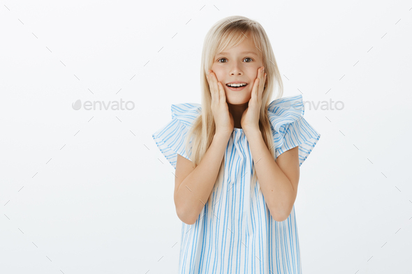 Adorable european young girl with blond hair in trendy blue blouse, gasping, holding hands near open