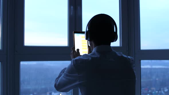 Young Businessman in Headphones Uses Tablet By the Panoramic Window in the Evening
