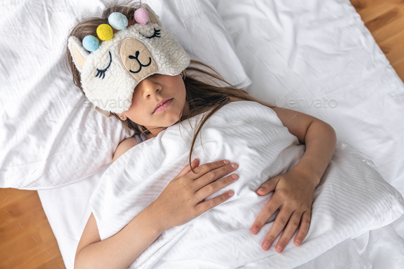 A little girl sleeps on a pillow wearing a funny sleep mask.
