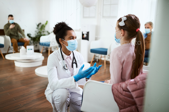 Black doctor with face mask talking to small girl in waiting room at the clinic.