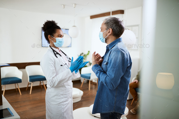 Happy patient and black female doctor wearing face masks while communicating in waiting room.
