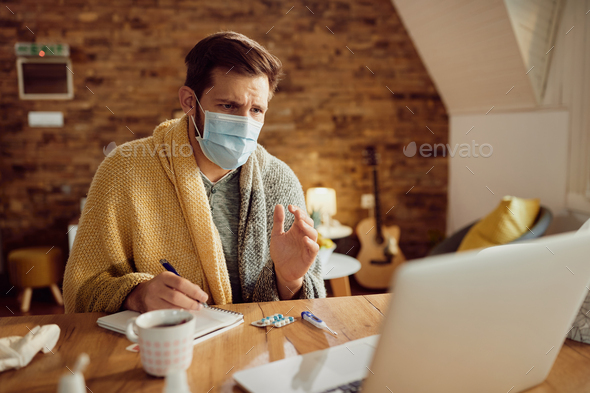 Ill man with face mask taking notes while having video call with doctor from home during coronavirus