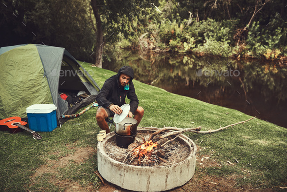 Cooking over the campfire. Shot of a young man preparing food at a campsite fire.