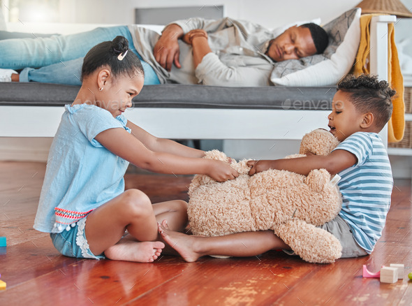 Shot of two siblings fighting over a teddy on the floor while their dad sleeps on the couch at home