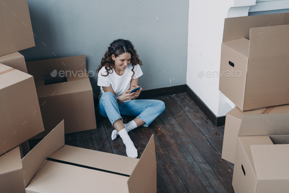 Modern girl sits with boxes uses smartphone app, choosing moving company and mover for relocation