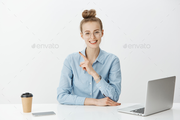 Positive good-looking female entrepreneur in glasses and blue-collar shirt, pointing at upper left c