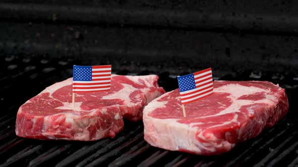 Two juicy rib-eye steaks sitting on the grill and cooking with two tiny American flags tooth picked alt