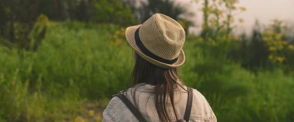 Behind shot of young girl with backpack and hat in the green field at sunset alt