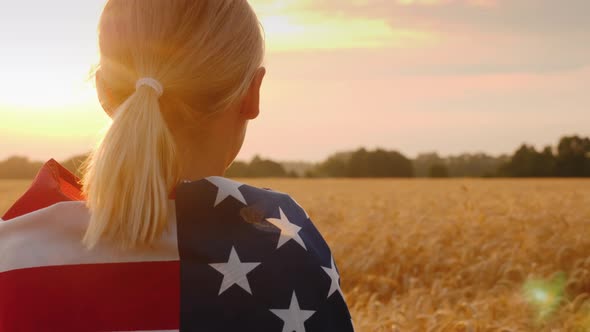 A Woman with a USA Flag on Her Shoulders Enjoys a Field of Wheat at Sunset alt