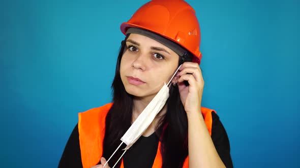 Female Construction Worker in Overalls Putting on Medical Mask on Face on Blue Background alt