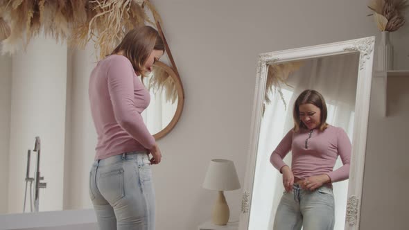 Unhappy Pretty Female Trying to Squeeze Into Old Tight Pair of Jeans in Front of Mirror alt
