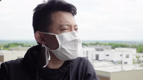 A Young Asian Man in a Face Mask Stands on a Balcony and Looks at a Cityscape Below alt