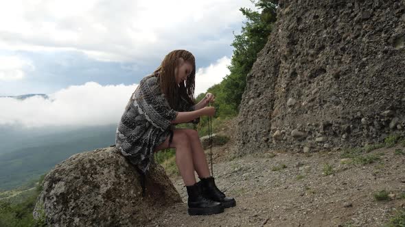 A Girl in a Gray Cape Sits on a Stone and Ties Her Shoelaces