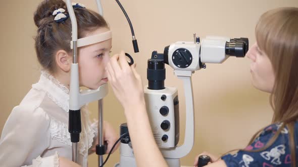 Woman Eye Doctor Is Correcting Head of Little Girl Near Biomicroscopy Apparatus alt