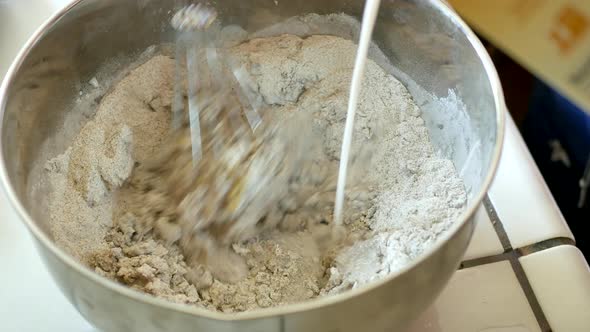 Mixing a very dry batter of wheat flower, eggs, oil and milk in a large metal mixing bowl with a whi alt
