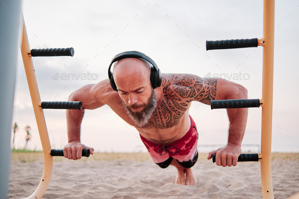 Athletic man using parallel bars to do push-ups while training calisthenics at the beach.