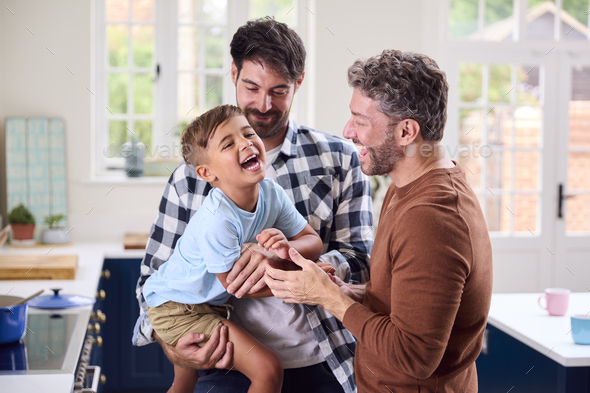 Same Sex Family With Two Dads Cooking In Kitchen With Son Sitting On Counter