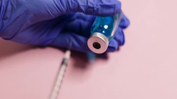 Womans Hand in Blue Latex Glove Holds Syringe Injection and Glass Bottle with Liquid Vaccine on Pink