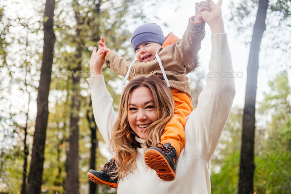 Smiling mother carrying son on shoulder and having fun in autumn park. iggyback ride outdoors