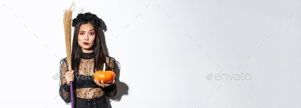Worried and confused asian woman in witch costume looking nervous, holding broom and pumpkin, trick