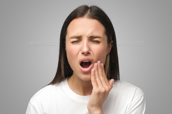 Studio shot of woman feeling pain, holding cheek suffering from bad tooth ache, isolated on gray