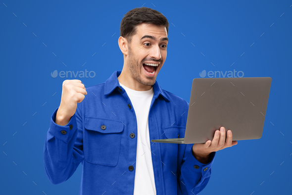 Excited man in blue shirt looking at laptop screen with WOW expression, isolated on blue