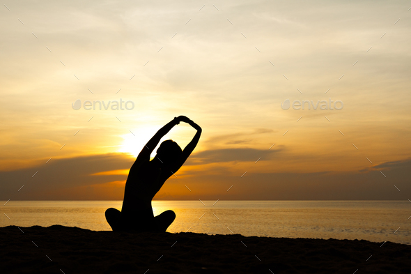Silhouette of stylish woman sit in lotus pose doing yoga and mediation on beach in sunrise