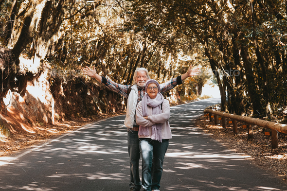 Senior couple walking in the road covered by trees shade. Elderly man and woman hike in the woods