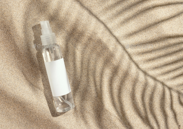 Spray bottle with blank label on beige sand top view, palm leaf hard shadow. Cosmetic mockup