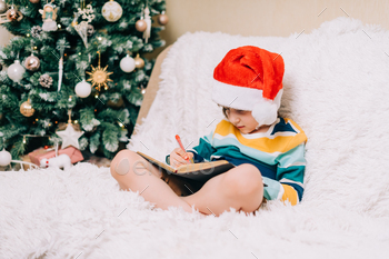Smiling Kid boy in red Santa hat sit on couch and writing the letter to dear Santa at home. Child