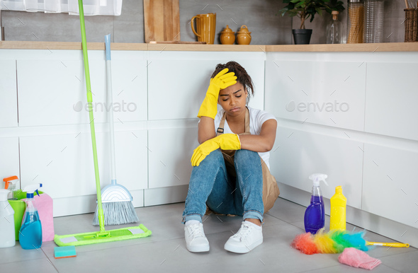 Despaired tired young black woman in rubber gloves sits on floor with mop, cleaning supplies suffers