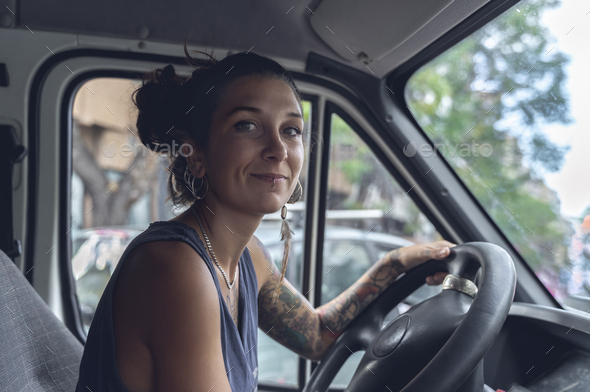Young woman, with tattoos and piercings, driving a van, looks at the room with satisfied expression.