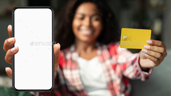 Happy millennial black female show credit card, smartphone with blank screen in living room interior