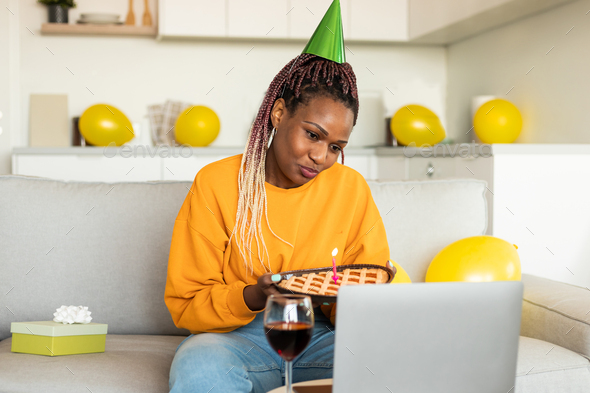 Online party. Sad single black woman in party hat celebrating b-day online, holding pie and looking