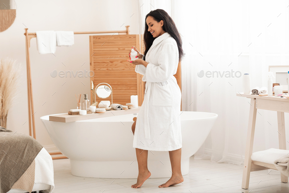 Black Woman Holding Body Balm Bottle Moisturizing Skin In Bathroom