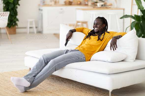 Smiling Young Black Man Resting On Couch With Remote Controller In Hand