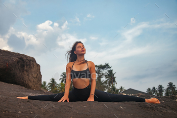 Young woman does yoga position splits or twine for healthy lifestyle on tropical coast, looking away