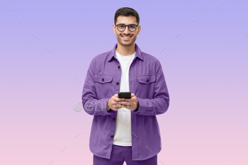 Smiling young man in casual purple shirt and glasses, holding smartphone, isolated on purple