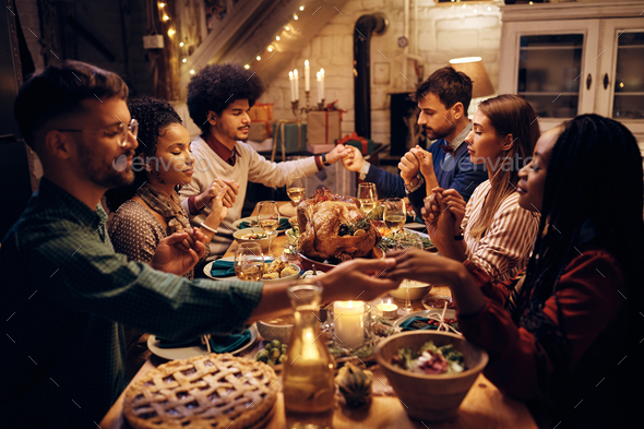 Multiracial group of friends saying grace during Thanksgiving dinner at dining table.