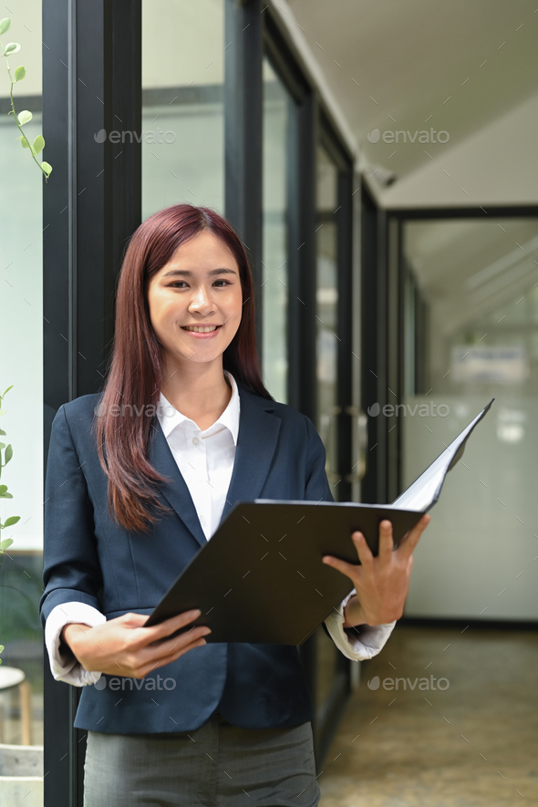 Asian businesswoman in formalwear holding folder and standing near glass window in office.