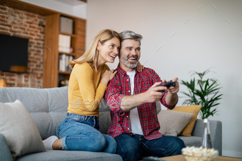 Happy mature european wife hugging husband with joystick, enjoy online game on computer, sit on sofa