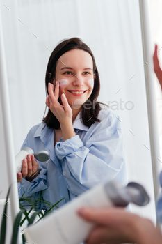 Smiling young woman applying cream on face looking in mirror indoors. Home beauty skincare concept