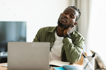 Tired black man suffering from neck spasm while working on laptop and rubbing his muscles, sitting