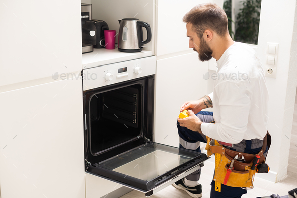 Repairman Examining Oven With Screwdriver In Kitchen With Tool Case