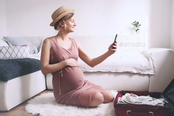 Pregnant woman packing suitcase for hospital or journey, making list or taking photo with smartphone