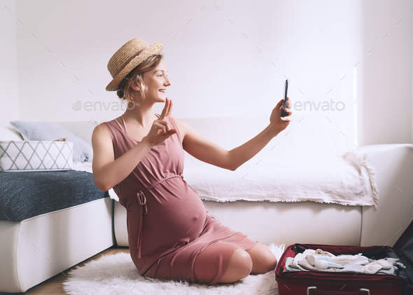 Pregnant woman packing suitcase for hospital or journey, making list or taking photo with smartphone